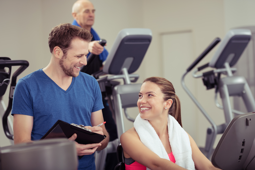 Attractive young woman with a trainer at the gym smiling up at him as they discuss her progress in a health and fitness concept