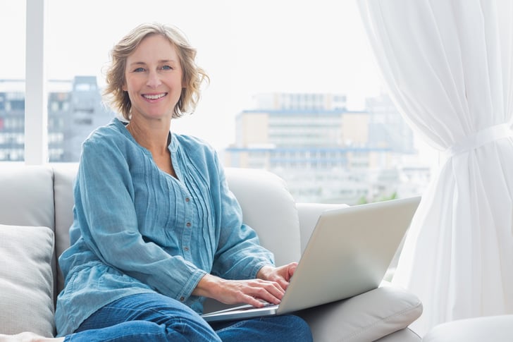 woman sitting on her couch using laptop smiling at camera at home in the sitting room